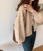 アプリコットのランタンスリーブショートカーディガンを着用し、白いTシャツと濃いデニムを合わせた女性のバストアップ。ボリューム袖とフロントのパッチポケットが特徴。