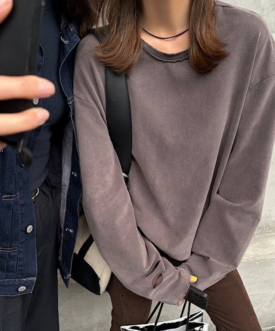 グレーブラウンのダメージ加工ニットを着用した女性のバストアップ。襟元のクラッシュ加工とレイヤードされた白Tシャツが確認できる。