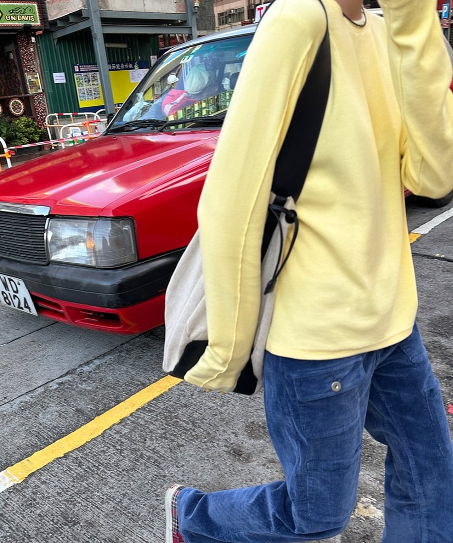 イエローのアウトシームロングTシャツとネイビーのコーデュロイパンツを着用した人物が、赤いタクシーの横を歩いている全身ショット。