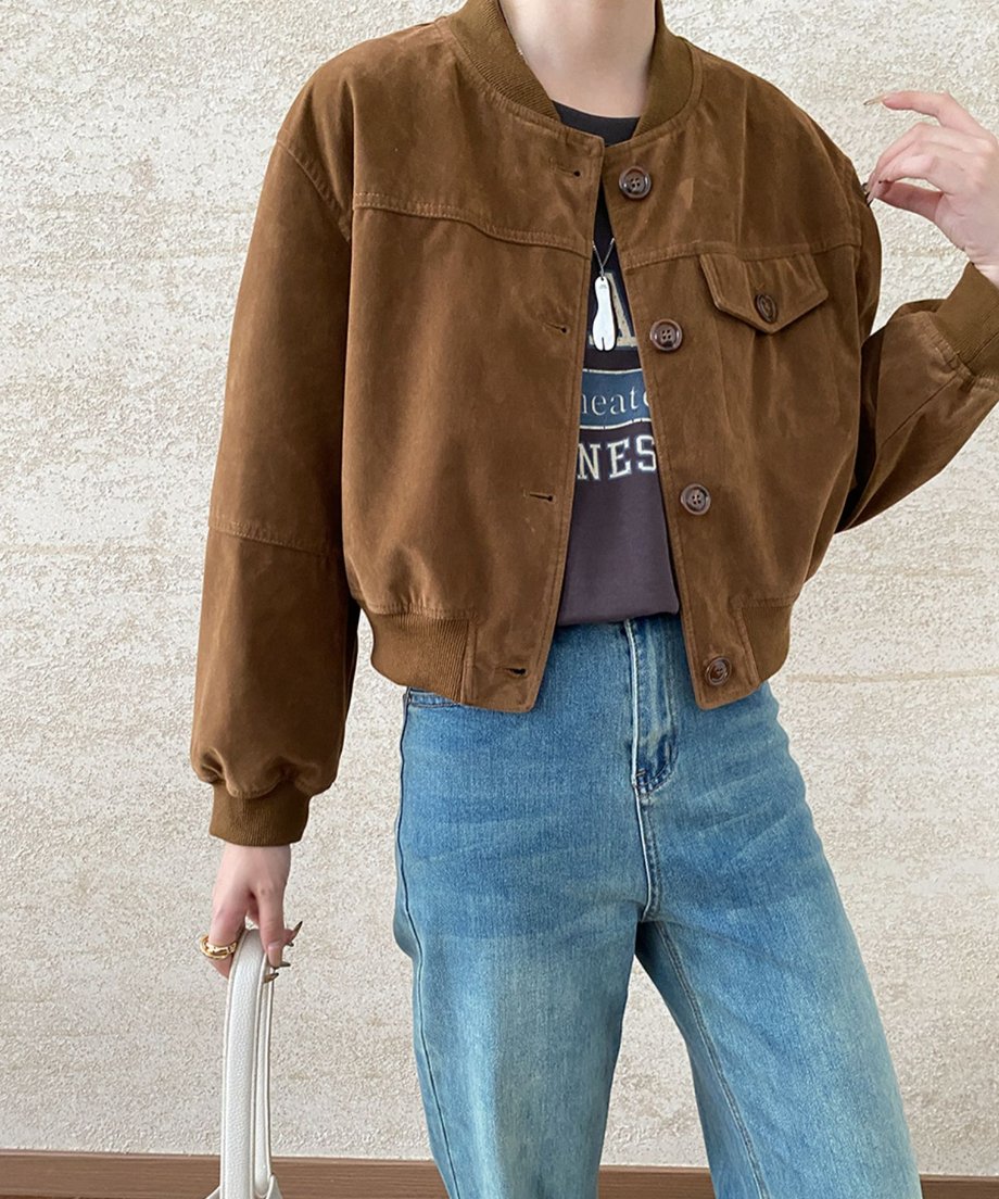 キャラメルのフェイクスエードジャケットのバストアップ。ロゴTシャツとデニムパンツを合わせ、白いバッグと指輪などのアクセサリーが見える。