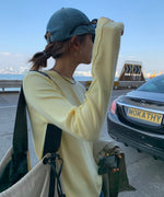 イエローのアウトシームロングTシャツを着て、デニムキャップを被り、車と海を背景に横向きに立っている人物のバストアップ。