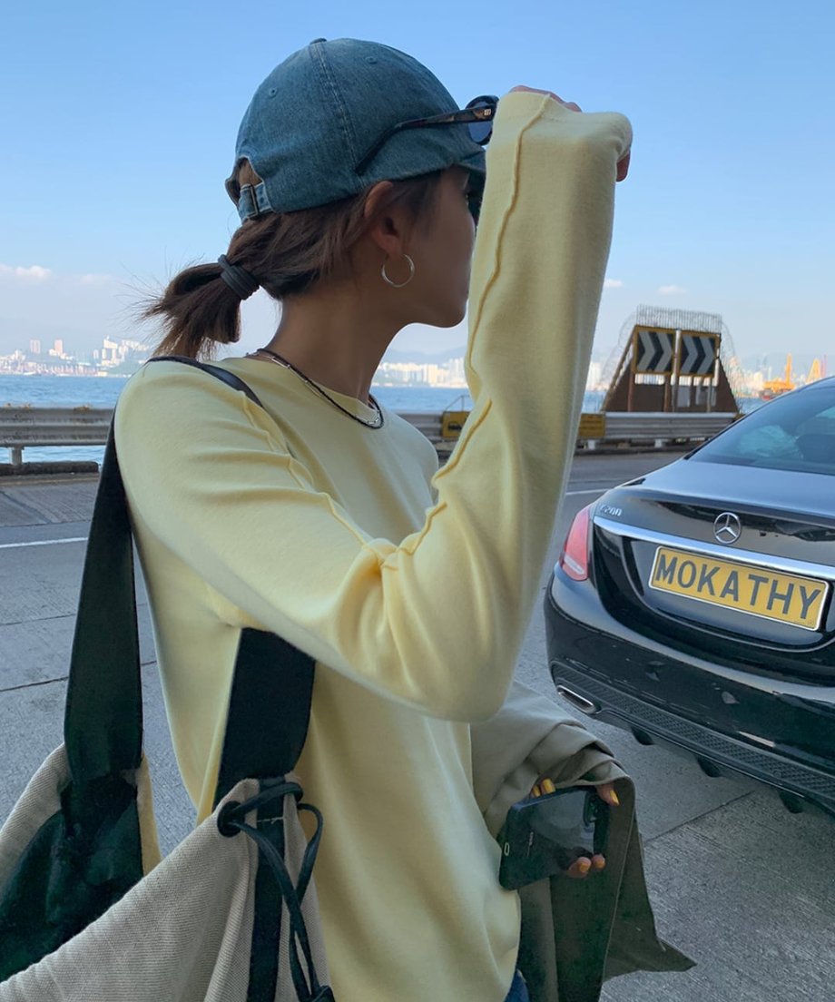 イエローのアウトシームロングTシャツを着て、デニムキャップを被り、車と海を背景に横向きに立っている人物のバストアップ。