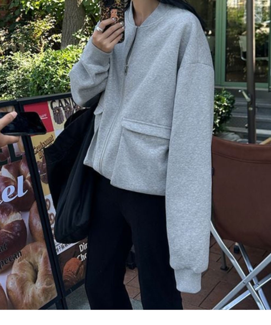グレーのラージポケットブルゾンを着用した女性が屋外に立っている写真。ポケットに手が入れられ、フロントの大きなフラップポケットが確認できる。