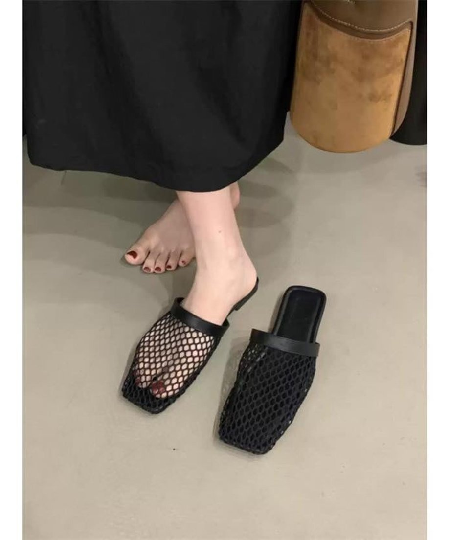 黒いロングスカートを着用した女性の足元。ブラックのスクエアトゥサンダルを片足だけ履いており、赤いペディキュアが見える。隣にもう一足置かれている。