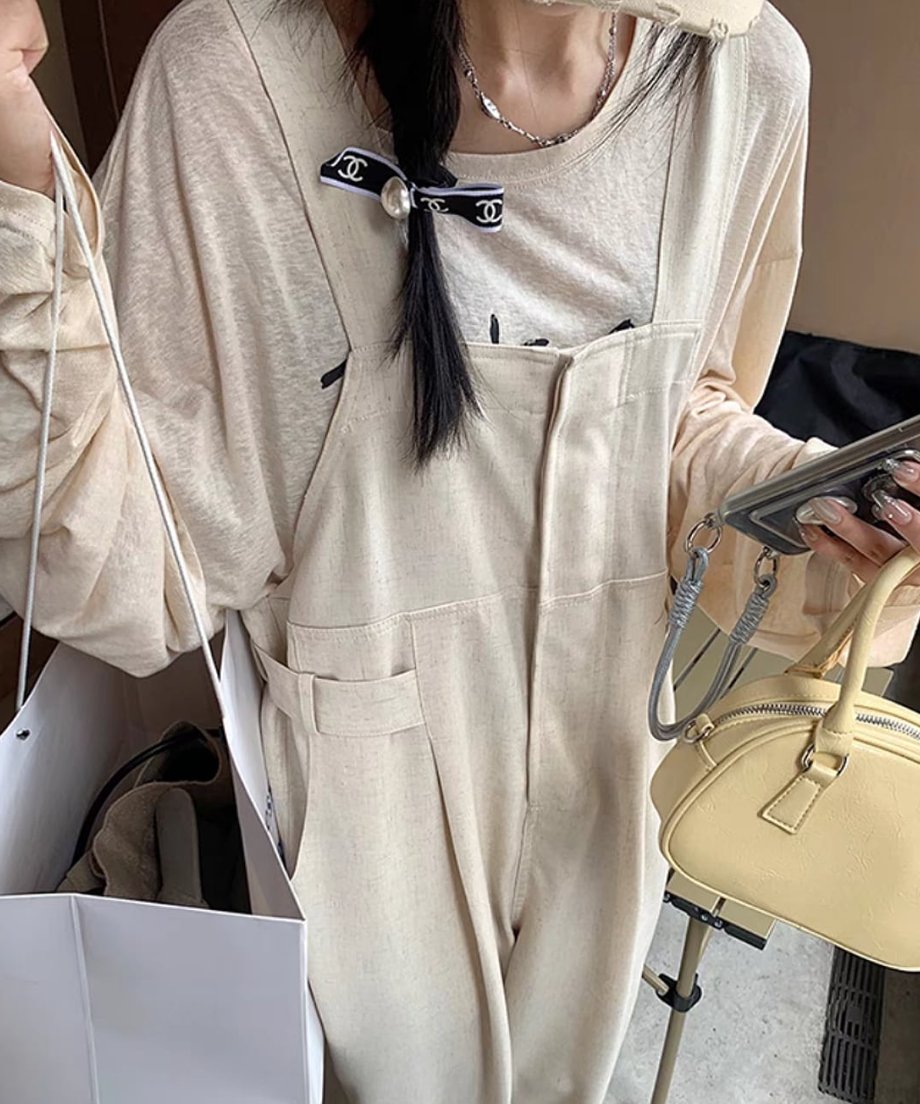 カフェのような場所で上半身を写した女性の写真。ベージュのオーバーオールに、オーバーサイズの長袖Tシャツをインナーに着用。黄色のミニバッグを持ち、三つ編みにリボンのヘアアクセサリーを付けている。