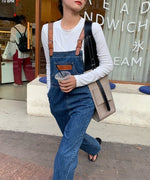 青いデニムオーバーオールを着用し、白の長袖Tシャツをインナーにした女性の全身ショット。茶色の革風ストラップがアクセントになっている。