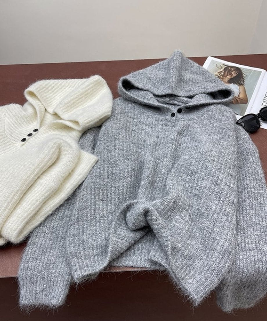 オフホワイトとグレーのフード付きニット2着が平置きされた画像。リブ編みと3つの黒いボタンのディテールが見える。