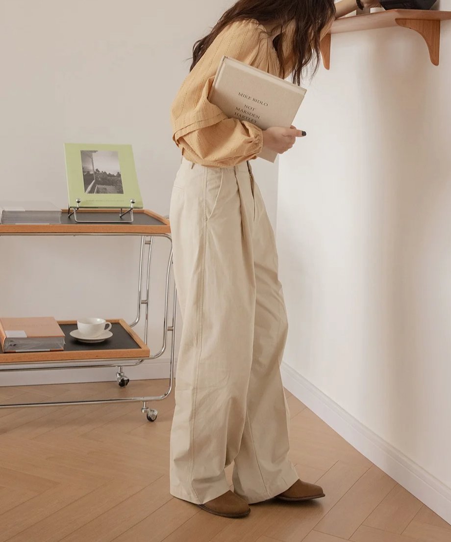 オフホワイトのワイドスラックスを着用し、キャメル色のブラウスと茶色のスリッパ風の靴を合わせた女性のサイドビュー。ノートとペンを持ち、室内の壁際に立っている。