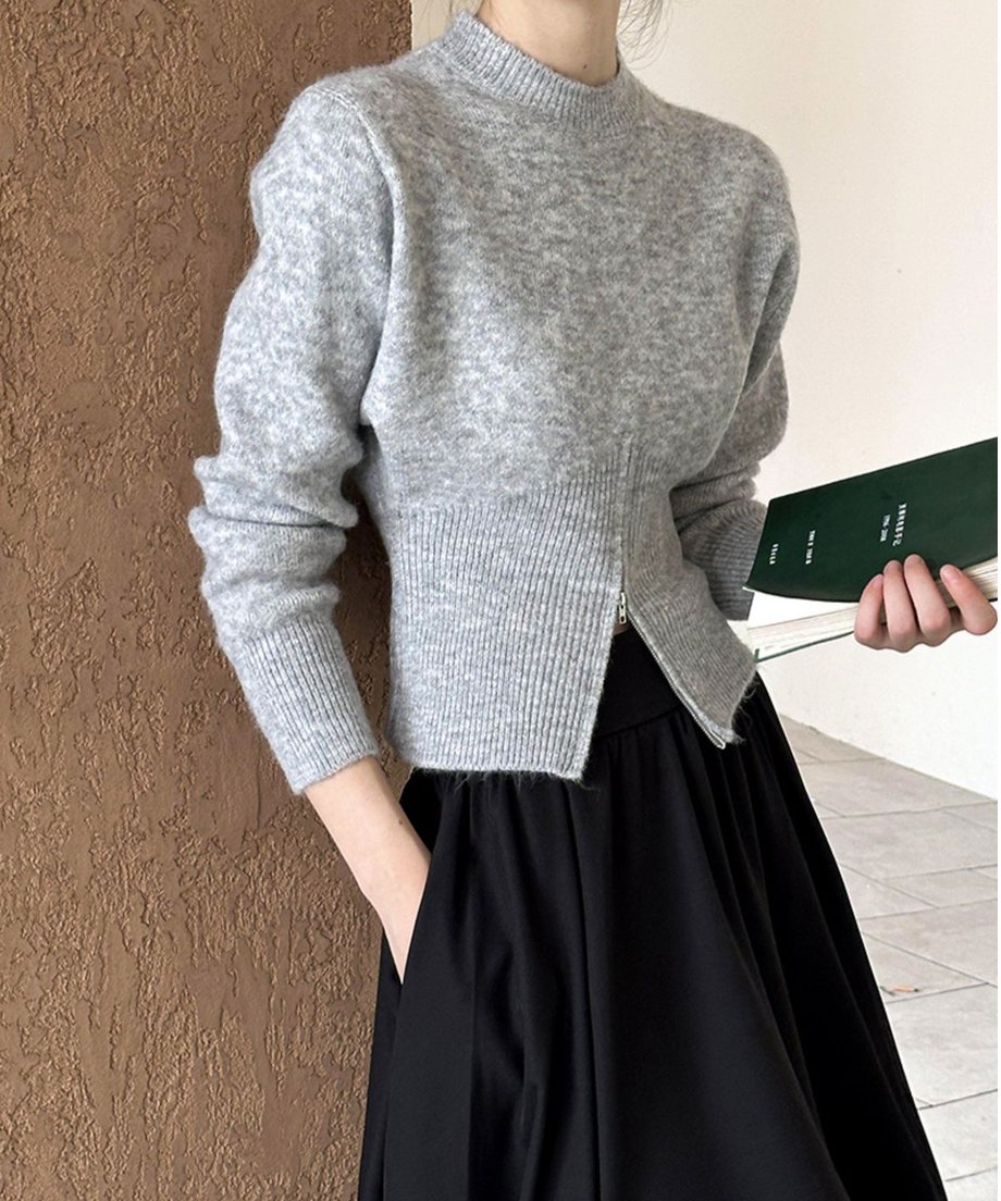 グレーのハーフジップニットと黒いロングスカートを着用した女性の半身像。左手をポケットに入れている。
