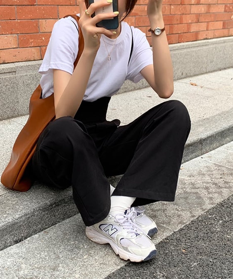 ブラックのハイウエストサスペンダーパンツと白Tシャツ、白ソックス、スニーカーを合わせた、縁に座る女性の足元とパンツ。