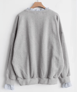グレーのレイヤードスウェットをハンガーにかけた背後の商品画像。ゆったりとしたシルエットと袖のスリット、ストライプの袖口フリルが見える。