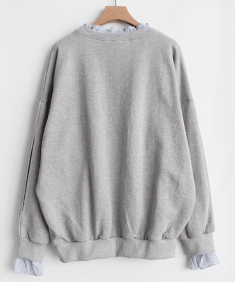 グレーのレイヤードスウェットをハンガーにかけた背後の商品画像。ゆったりとしたシルエットと袖のスリット、ストライプの袖口フリルが見える。