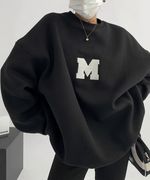 黒のオーバーサイズスウェットを着用し、マスクをつけている上半身。白い大きな「M」のアップリケが見える。