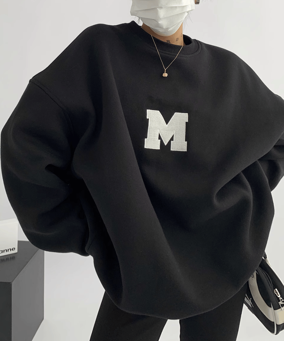 黒のオーバーサイズスウェットを着用し、マスクをつけている上半身。白い大きな「M」のアップリケが見える。