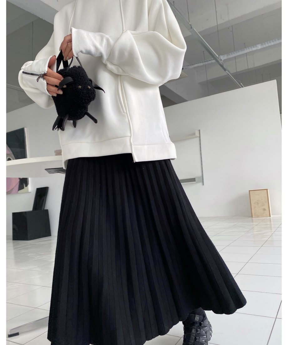 ホワイトのボリュームスウェットと黒のロングプリーツスカートを着用し、黒いファーのミニバッグを持った女性の全身。カジュアルなスニーカーと合わせたコーディネート。