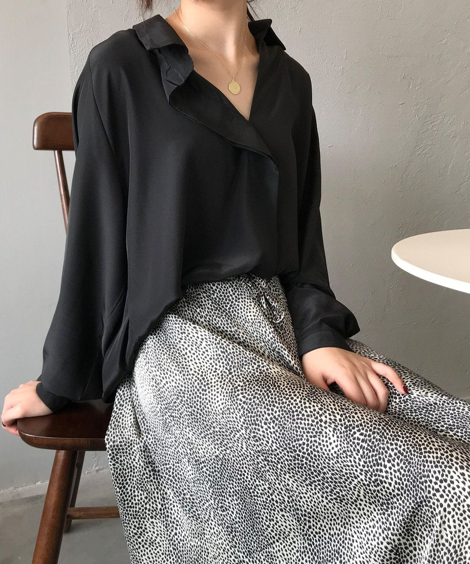 ランダムドット柄のロングテールスカートに黒のオーバーサイズシャツを着た女性が、木製の椅子に座っている上半身。