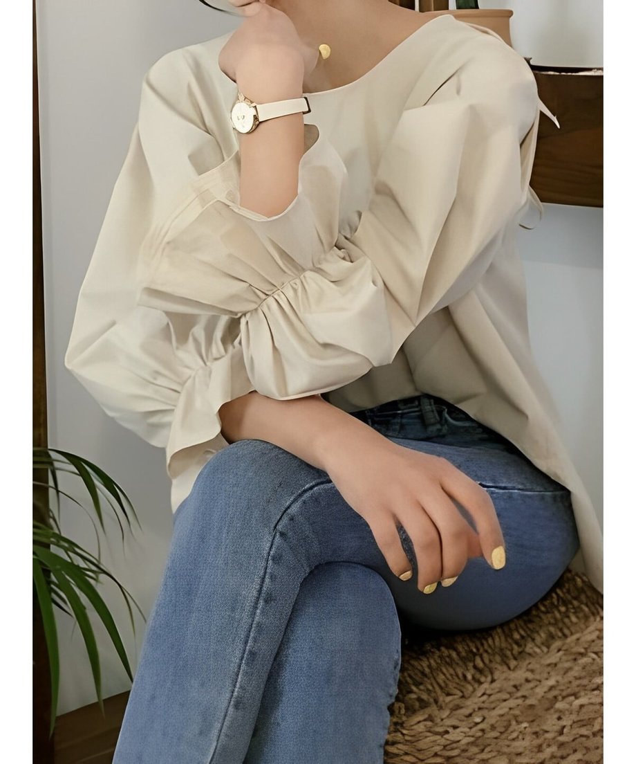 アプリコットのボリュームスリーブブラウスを着用した女性の手元クローズアップ。袖口はギャザーで絞られたフリル状になっており、白い時計と華奢なゴールドネックレスを身に着けている
