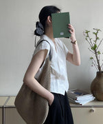 ホワイトのVネックブラウスと黒いボトムスを着用した女性の横顔。茶色いメッシュバッグを肩にかけ、手に持った本で顔の一部を隠している。