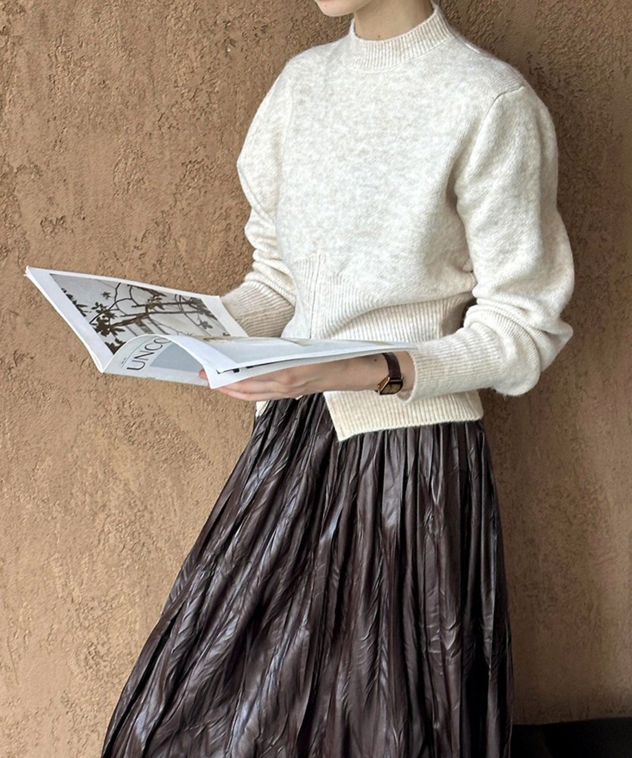 ベージュのハーフジップニットと光沢のある茶色いロングスカートを着用した女性の半身像。雑誌を手に持っている。