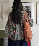 花柄のブラウスを着用し、茶色のトートバッグを肩にかけた女性の後ろ姿の画像。ボリューム袖のシルエットが確認できる。