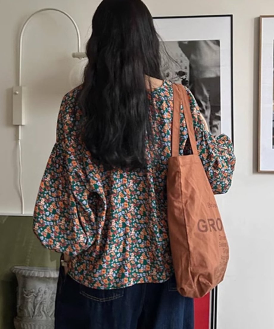 花柄のブラウスを着用し、茶色のトートバッグを肩にかけた女性の後ろ姿の画像。ボリューム袖のシルエットが確認できる。