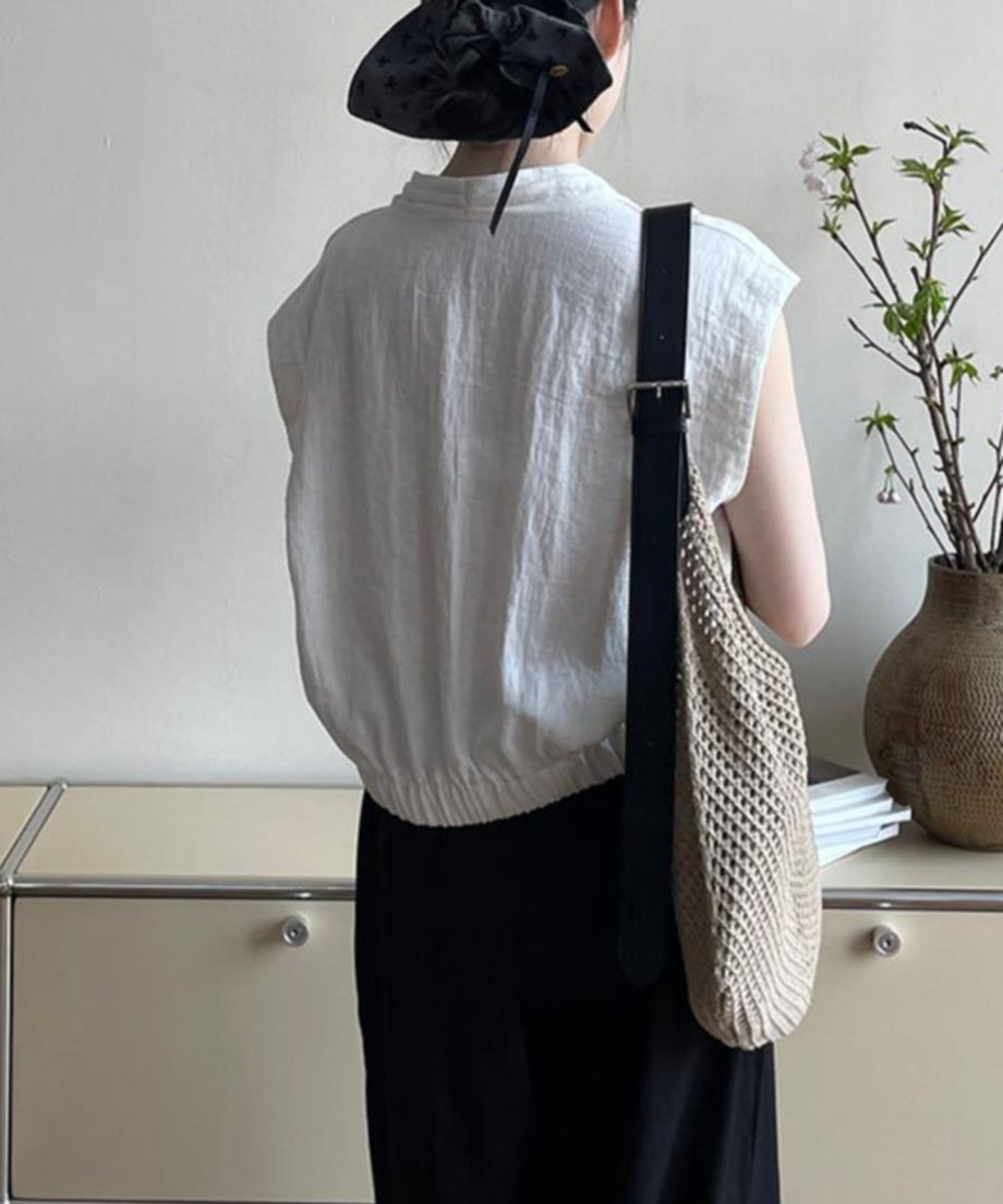 ホワイトのVネックブラウスと黒いボトムスを着用した女性の背面寄りアングル。肩に幅広のレザーストラップが付いたメッシュバッグをかけている。