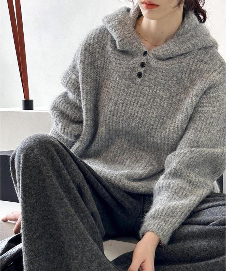グレーのフード付きニットとグレーのワイドパンツを着用した女性のクローズアップ。ニットはリブ編みで、フードの付け根に3つの黒いボタンが見える。