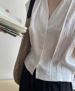 ホワイトのVネックブラウスのバスト付近をアップ。女性が手に持った本とメッシュバッグが写っており、ブラウスの前ボタンとVネックのディテールが確認できる。