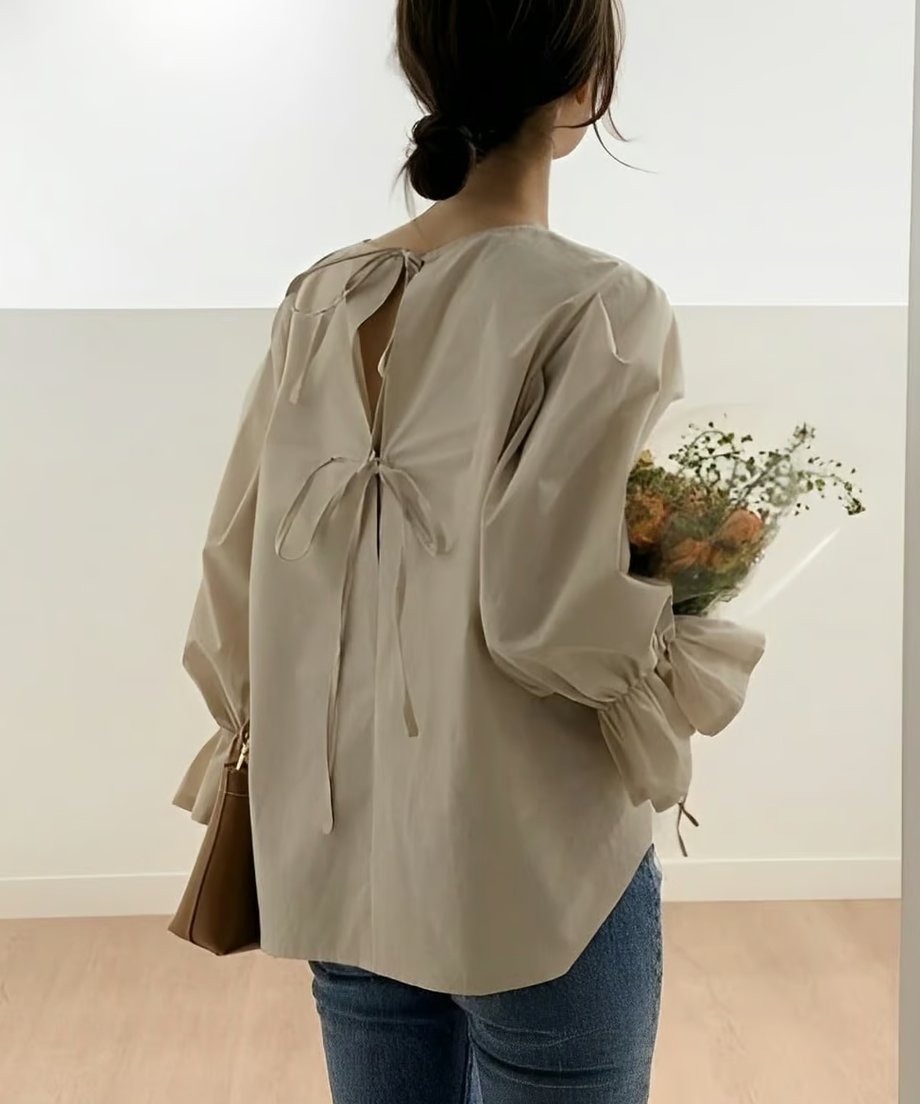 背中側を向いた女性で、ブラウスの背中のU字に開いたスリットと、結ばれた共布のリボンが特徴的。女性は茶色のバッグを持ち、花束を抱えている。