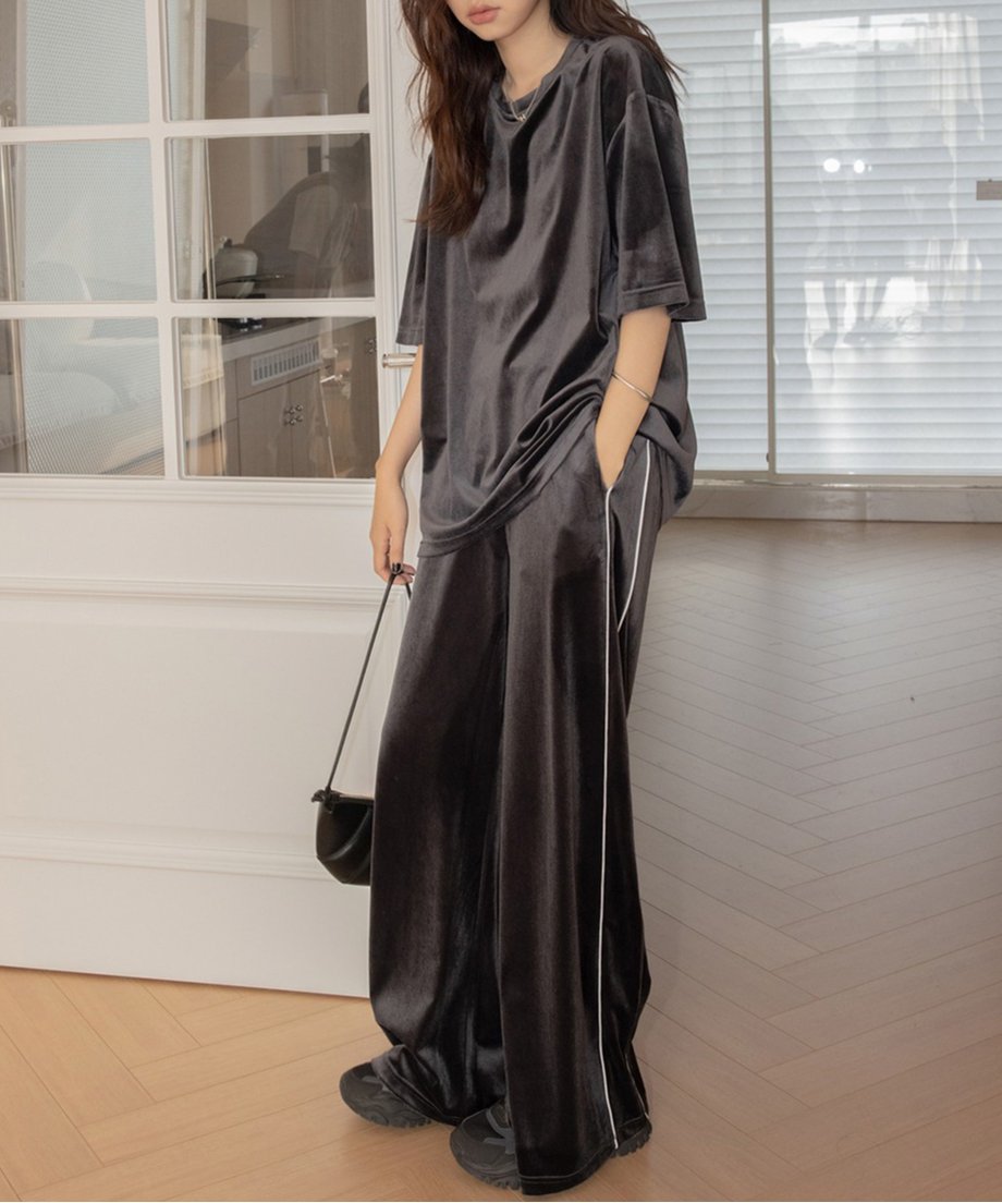 ダークグレーのベロアセットアップを着用した女性の全身。オーバーサイズTシャツとサイドに白いパイピングが入ったワイドパンツ。黒いショルダーバッグを持っている。