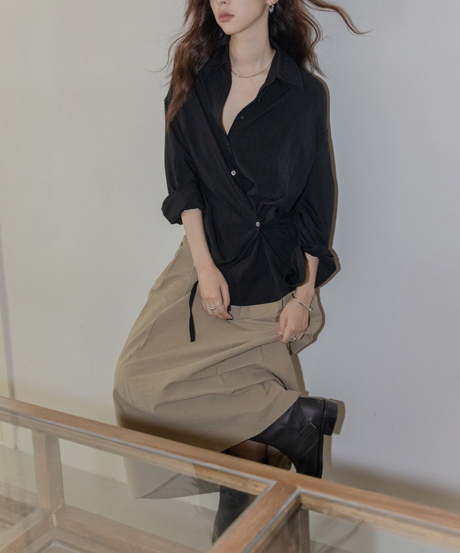 ブラックシャツとベージュのプリーツスカートの女性がポーズをとっている。スタイリッシュなファッション。