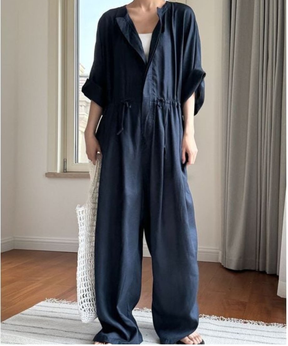 ネイビーのドローストのジャンプスーツを着用した女性の全身像。ワイドシルエットのパンツ部分と、ウエストのドローストリングが特徴。白いメッシュバッグを持っている。