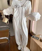 カフェのような屋内空間で、ベージュのワイドオーバーオールを着用した女性の全身写真。白のロゴ入り長袖Tシャツをインナーにし、ベージュ系のミニバッグを持っている。