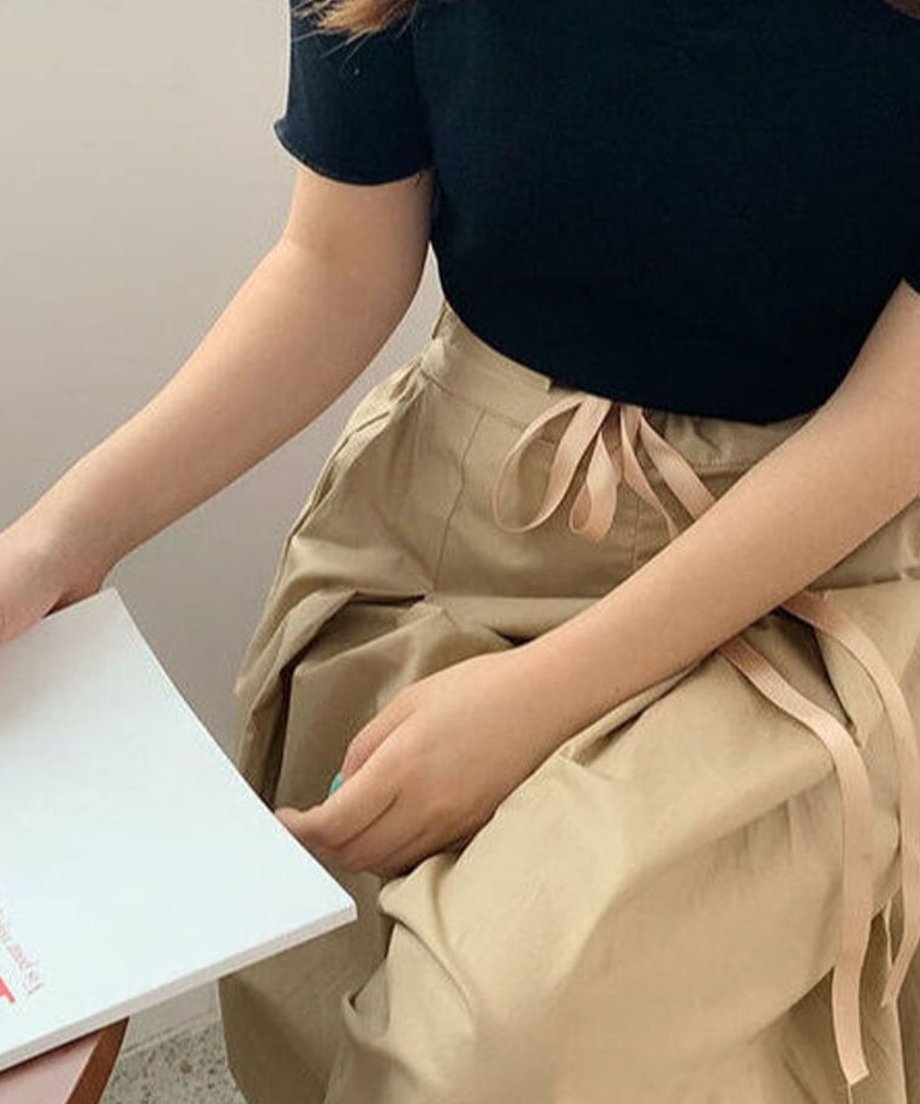 ベージュのフレアパンツのウエストと手元のクローズアップ。黒いトップスをインしており、光沢のあるベージュのリボンベルトが見える。