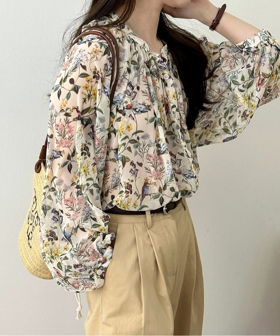 花柄ブラウスとベージュパンツのコーデ。 ストローバッグを持ち、髪に手を添えた女性の立ち姿。