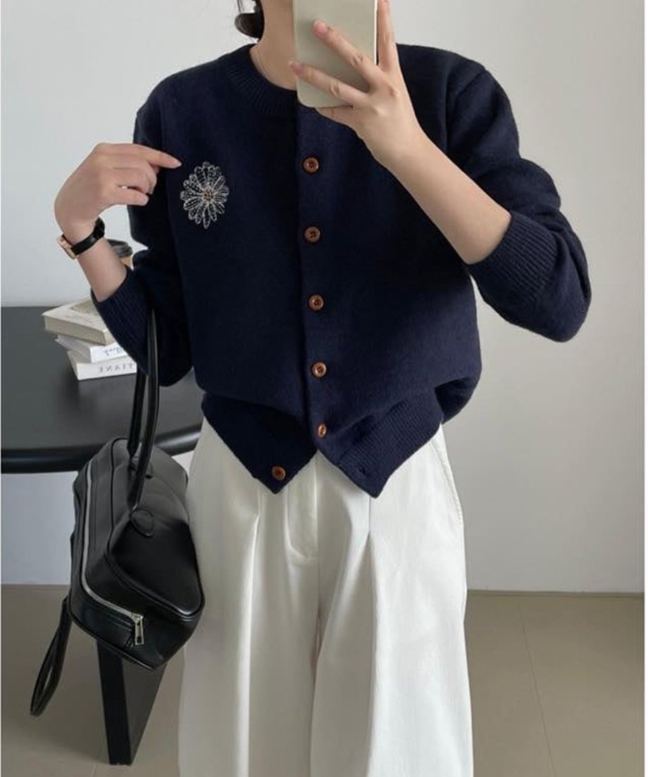 ネイビーのニットカーディガンを着用した女性の半身像。左胸に白い刺繍の花柄モチーフがあり、白のワイドパンツと黒のショルダーバッグを合わせている。