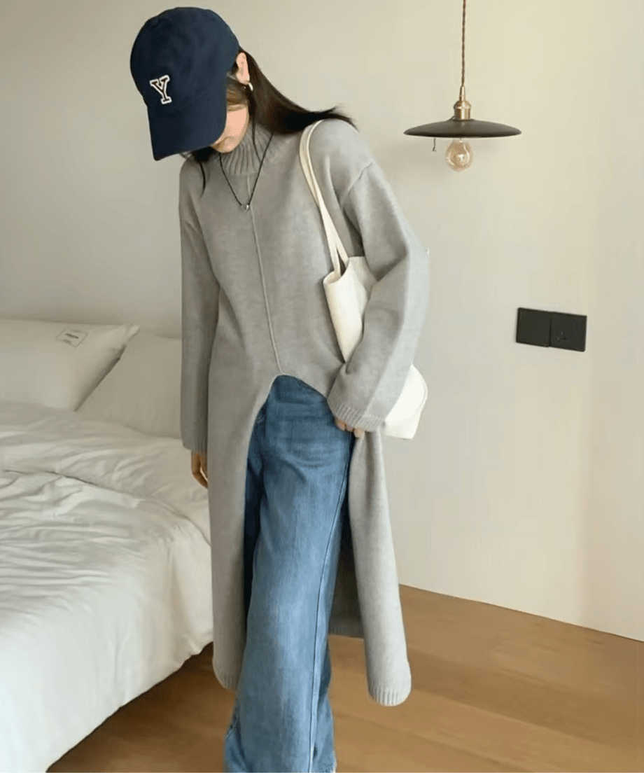 ライトグレーのタートルネックロングニットを着用した女性が、青いデニムパンツをレイヤードし、鏡の前でキャップとトートバッグを合わせたカジュアルスタイル。