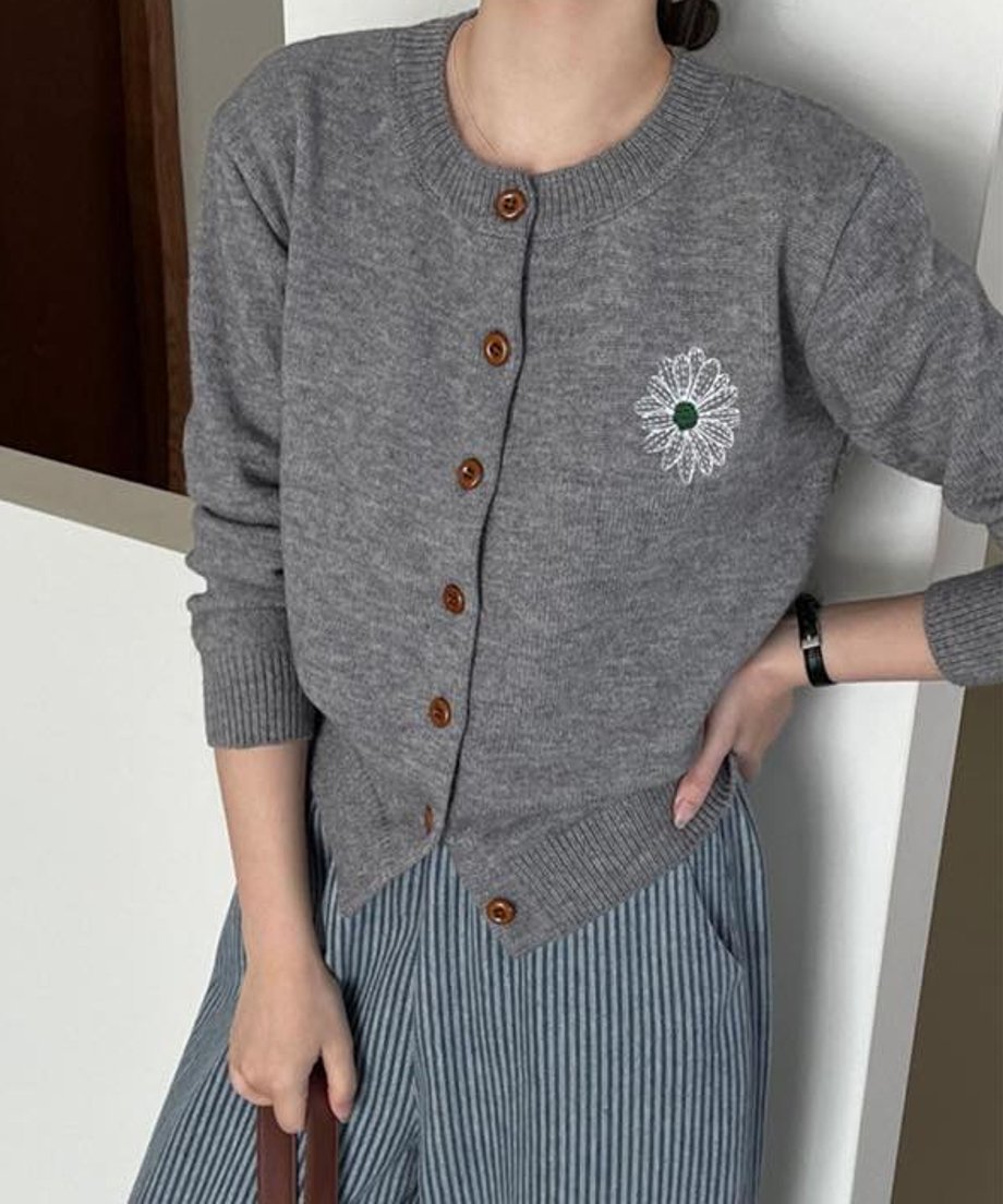 グレーの花柄刺繡ニットカーディガンを着用した女性のクローズアップ。白い花柄刺繍の中心は緑色。下には青と白のストライプ柄のボトムスを着用。