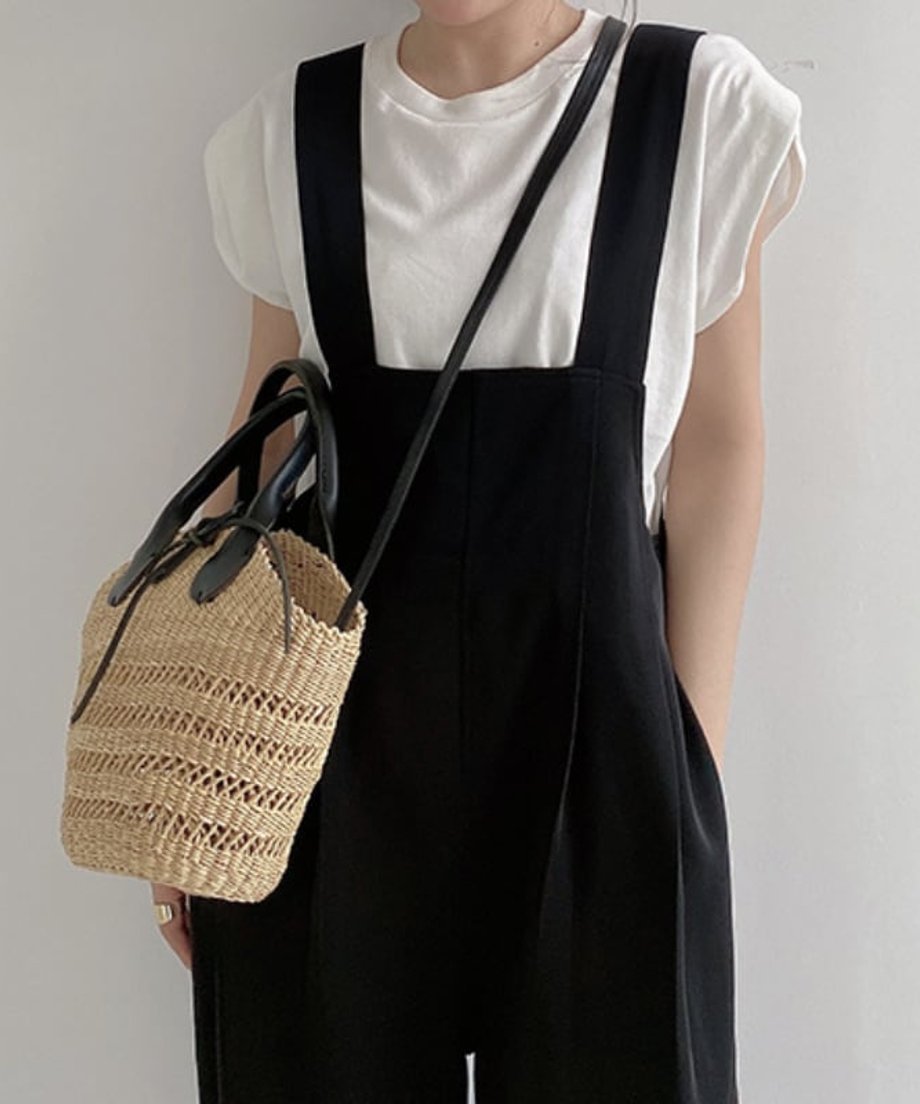 白いTシャツと黒いワイドレッグサロペットを着用し、ストロー素材のミニバッグを持った女性の上半身のクローズアップ。