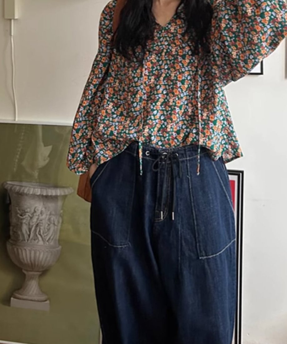 花柄のブラウスと濃紺のドローコード付きワイドデニムパンツを着用した女性が、片手を上げたポーズのコーディネート画像。