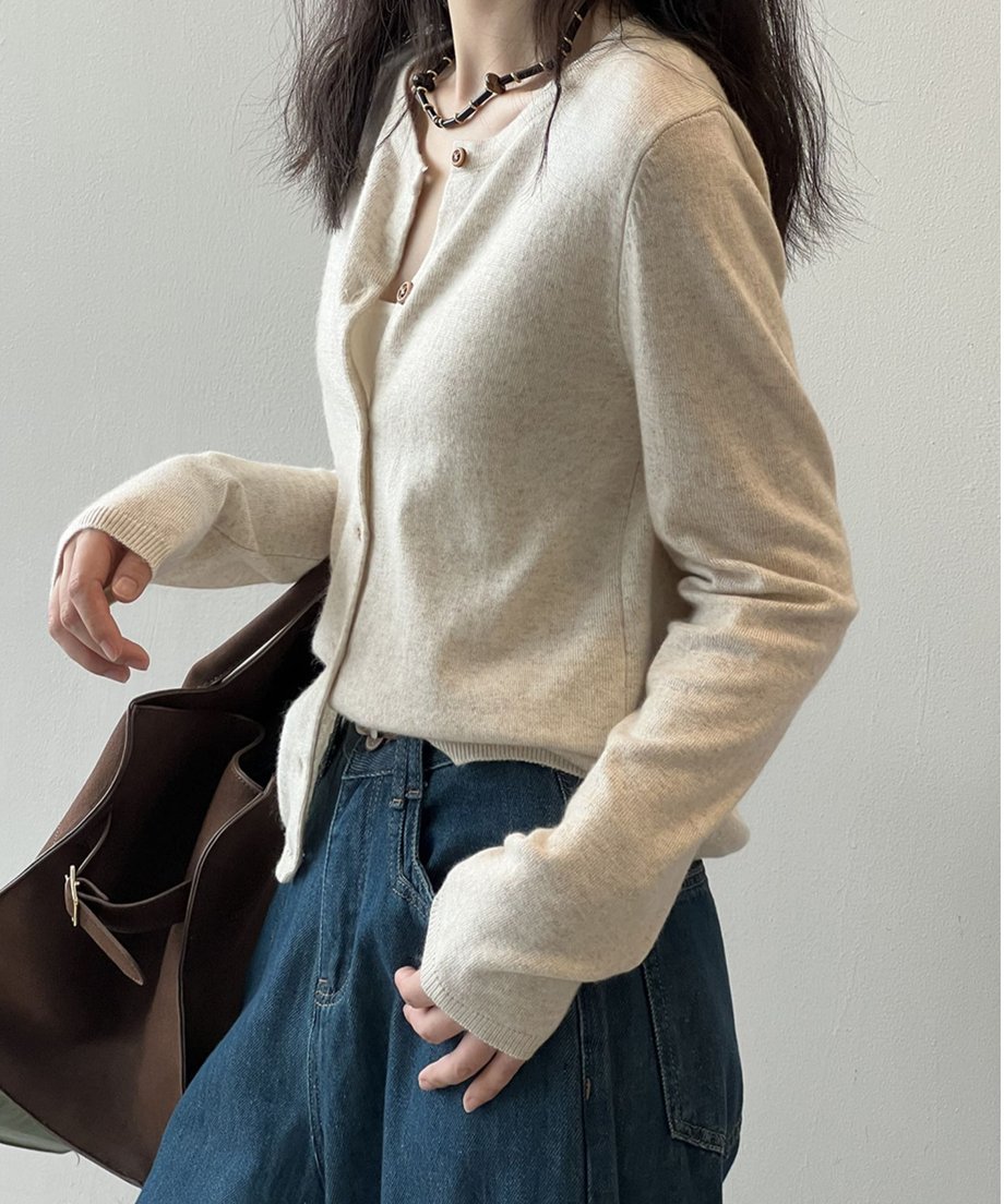 アプリコットのクルーネックカーディガンを着用した女性の横向きの半身像。ネイビーのワイドデニムに、茶色のレザートートバッグを合わせて着用している。