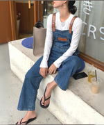デニムオーバーオールに白の長袖Tシャツを合わせた女性が、屋外のベンチに座ってリラックスしている全身ショット。
