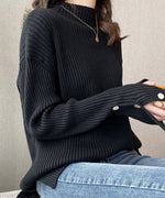 ブラックのモックネックニットを着用し、デニムを合わせて座っている女性のバストアップ。袖口の白いボタンとゴールドのネックレスが見える。