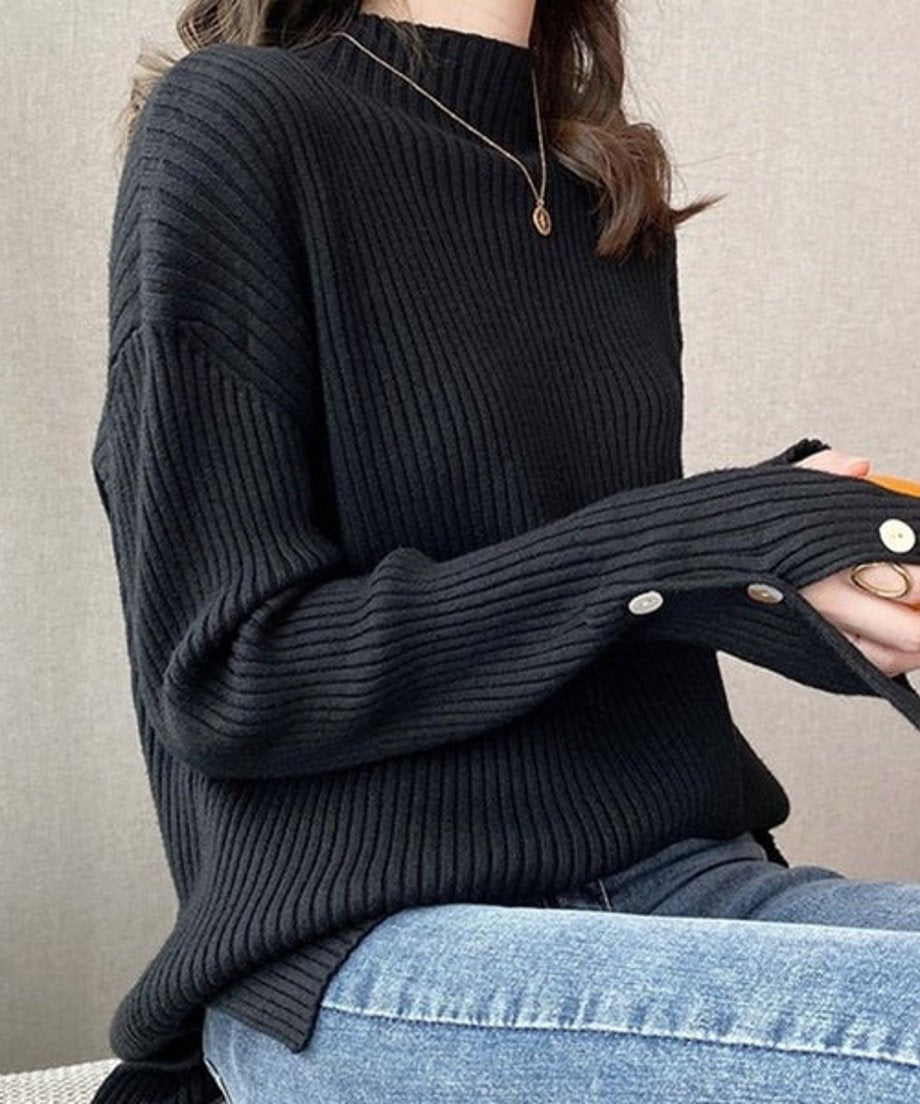 ブラックのモックネックニットを着用し、デニムを合わせて座っている女性のバストアップ。袖口の白いボタンとゴールドのネックレスが見える。