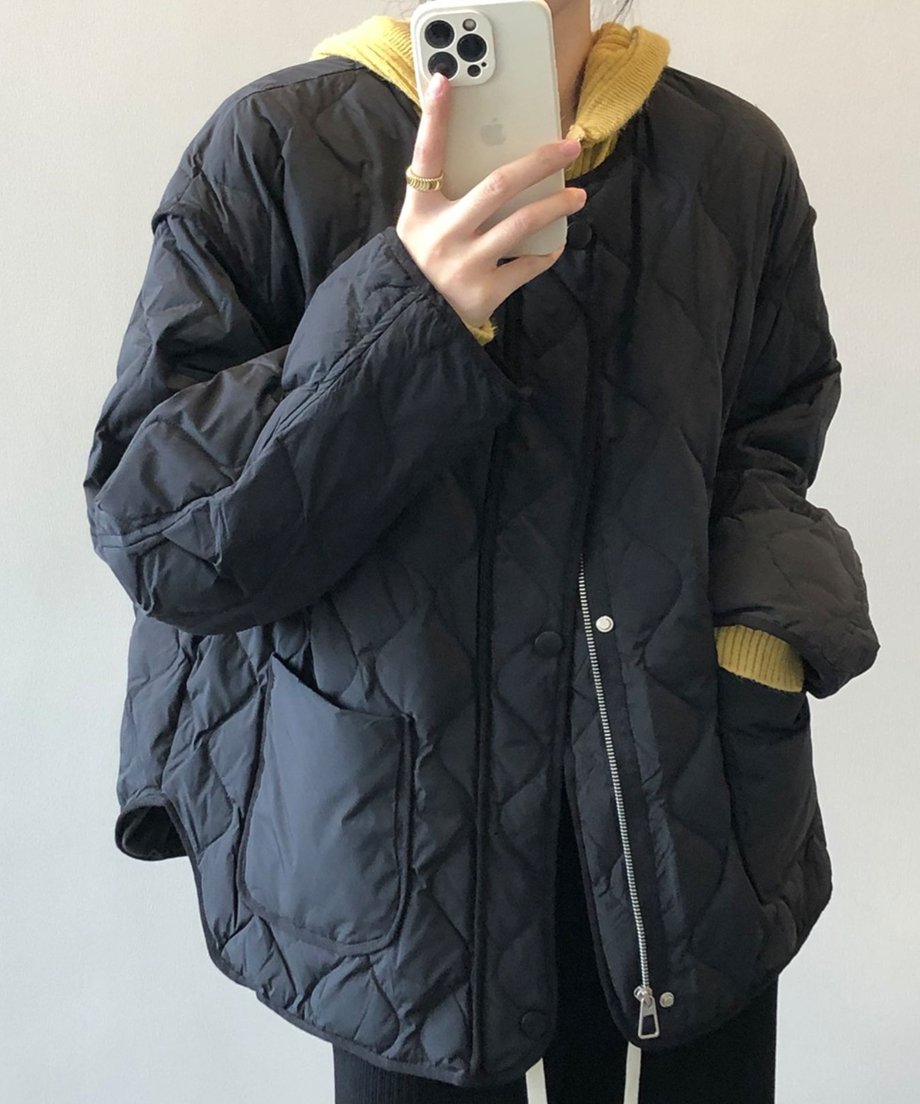 スマートフォンで自撮りをする女性が着用している、黒のノーカラーキルティングジャケットの上半身アップ。インナーに黄色のニットパーカーを合わせている。