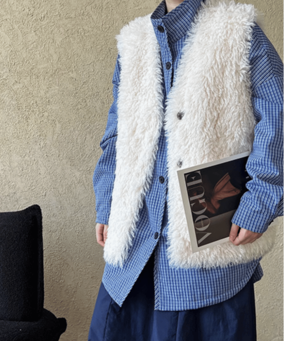 ホワイトのファーベストをブルーのチェック柄シャツにレイヤードし、雑誌を抱えている女性の画像。カジュアルな着こなし提案。