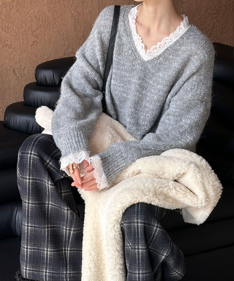 グレーのレース付きニットを着用し、チェック柄のパンツと合わせたコーディネート。片手に白いボアアウターと黒いハンドバッグを持った女性が座っている。