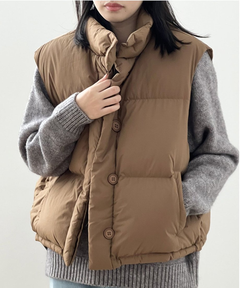 コーヒー色のベストを着用し、胸元に手を添えている女性の正面アップ。グレーのニットとの色合わせや、マットなボタンの質感が分かる。