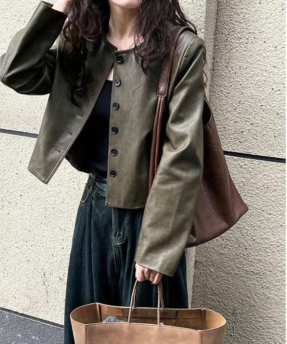 オリーブのレザージャケットを羽織り、黒のトップスとワイドデニムを着て、茶色のバッグを2つ持った人物の屋外での全身ショット。