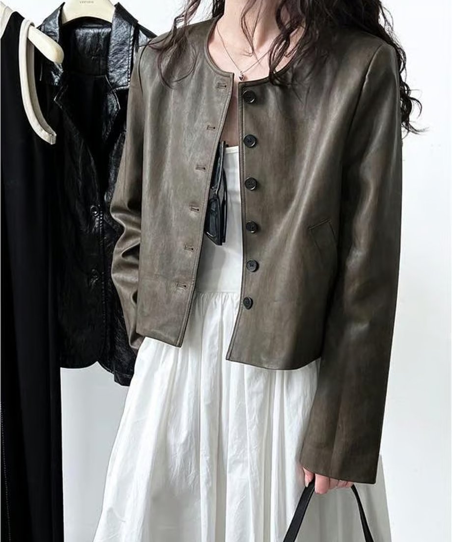 オリーブのノーカラーレザージャケットを白のワンピースに合わせ、ハンガーラックの服の前に立っている人物の姿。